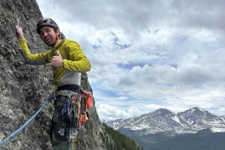 Man in green long sleeve and grey pants rock climbing in Rocky Mountain National Park with mountain range in the background.